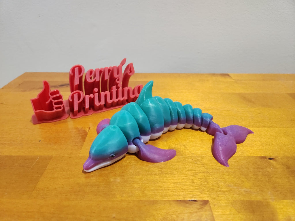 3D printed dolphin figurine in blue and purple colors on wood table with Perry's Printing sign
