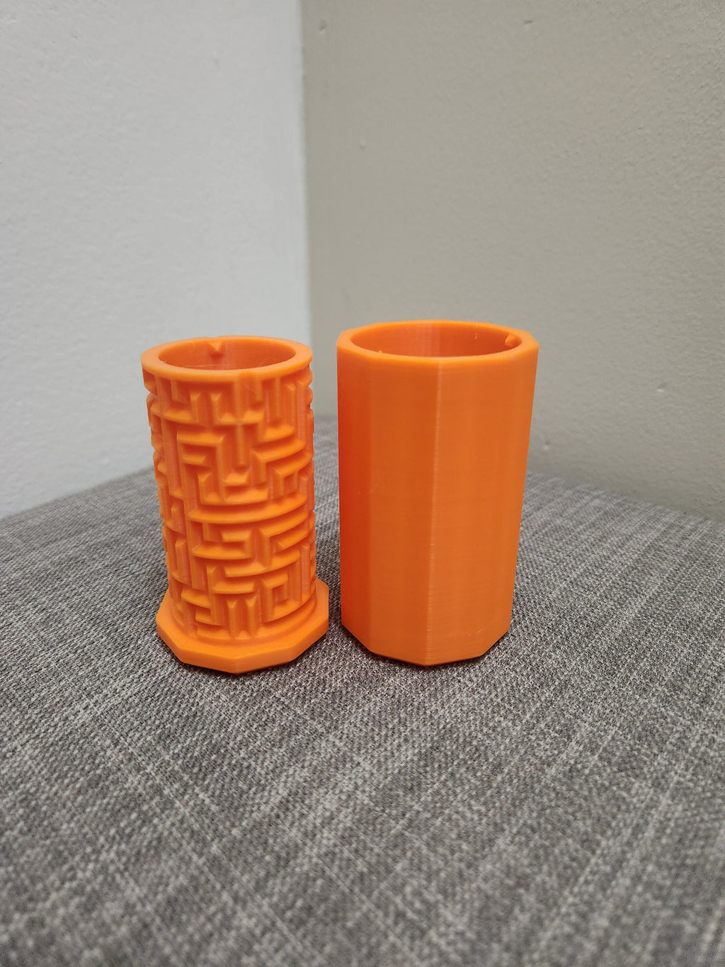 Two orange 3D printed cylindrical containers, one with a maze pattern, on a gray fabric surface.