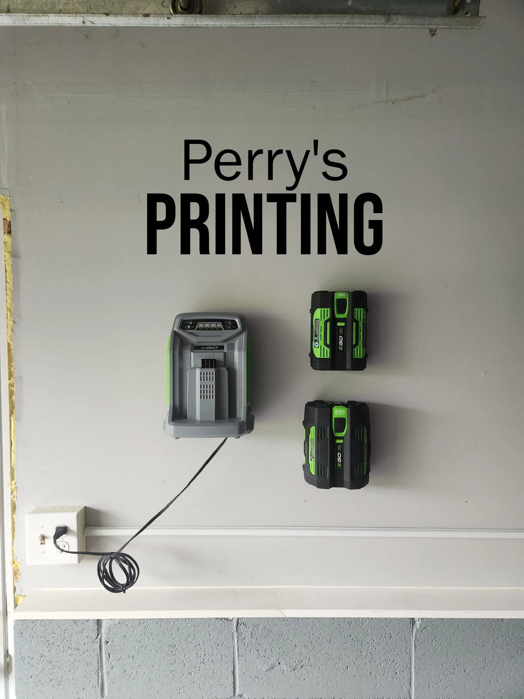 Wall-mounted green and black battery charger and batteries below 'Perry's PRINTING' sign.