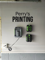 Wall-mounted green and black battery charger and batteries below 'Perry's PRINTING' sign.