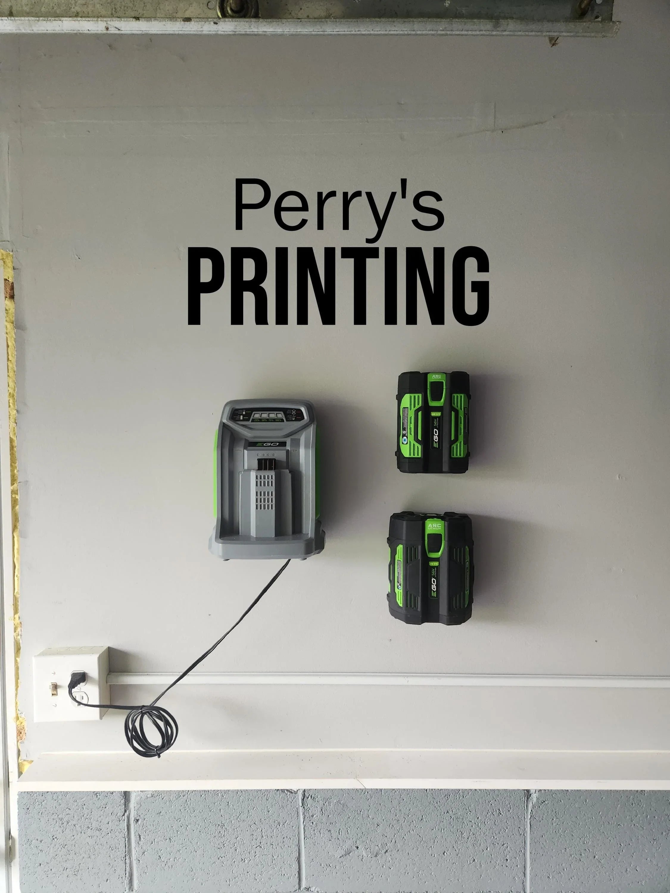 Wall-mounted green and black battery charger and batteries below 'Perry's PRINTING' sign.