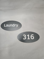 Brushed metal oval door signs with white text reading 'Laundry' and '316' on a light background.