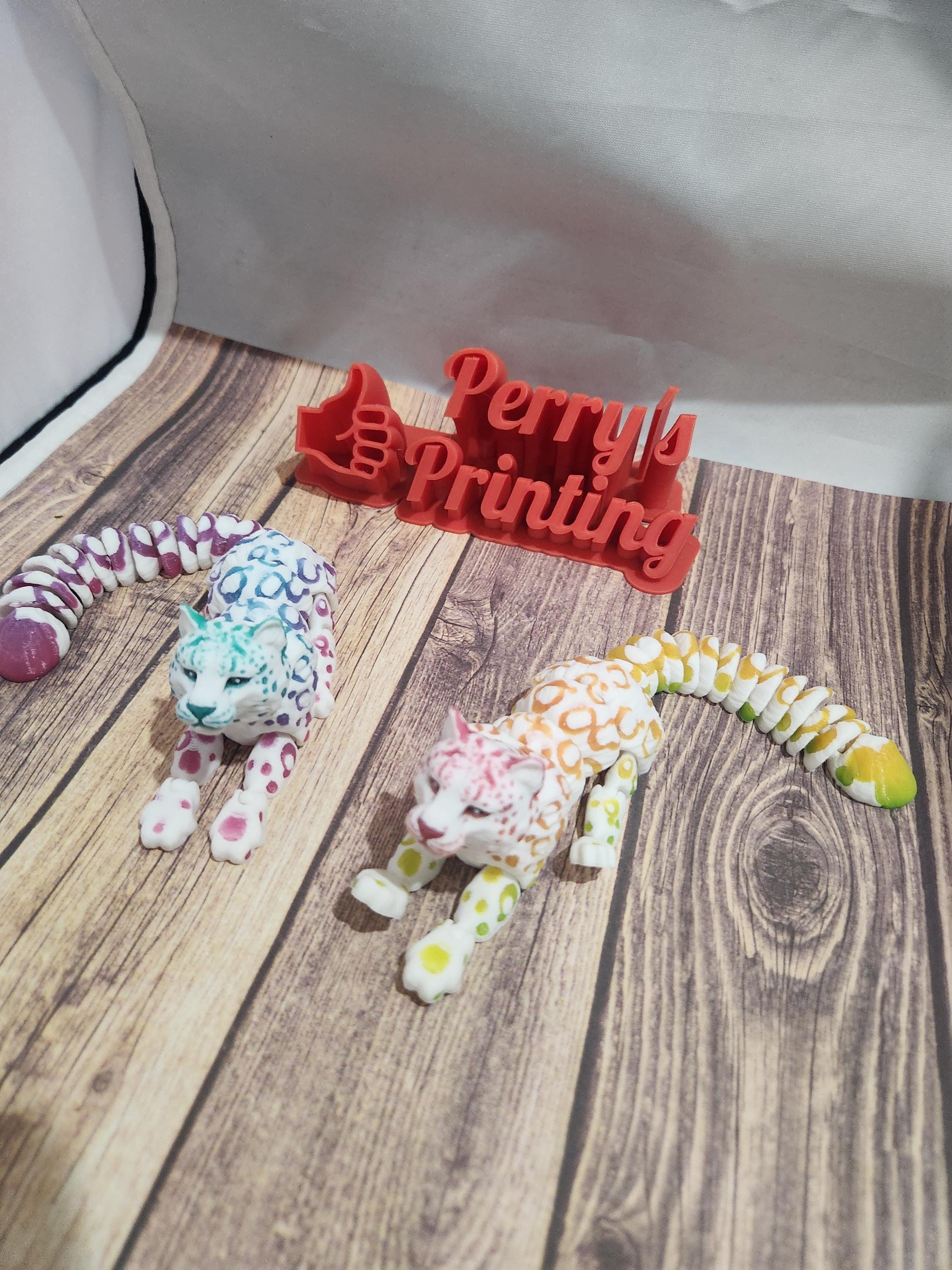 Two colorful 3D printed articulated leopard toys on wood surface, red Perry's Printing sign in back