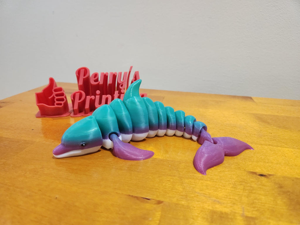Colorful 3D printed articulated dolphin toy on wood table with red printed sign and thumbs up.