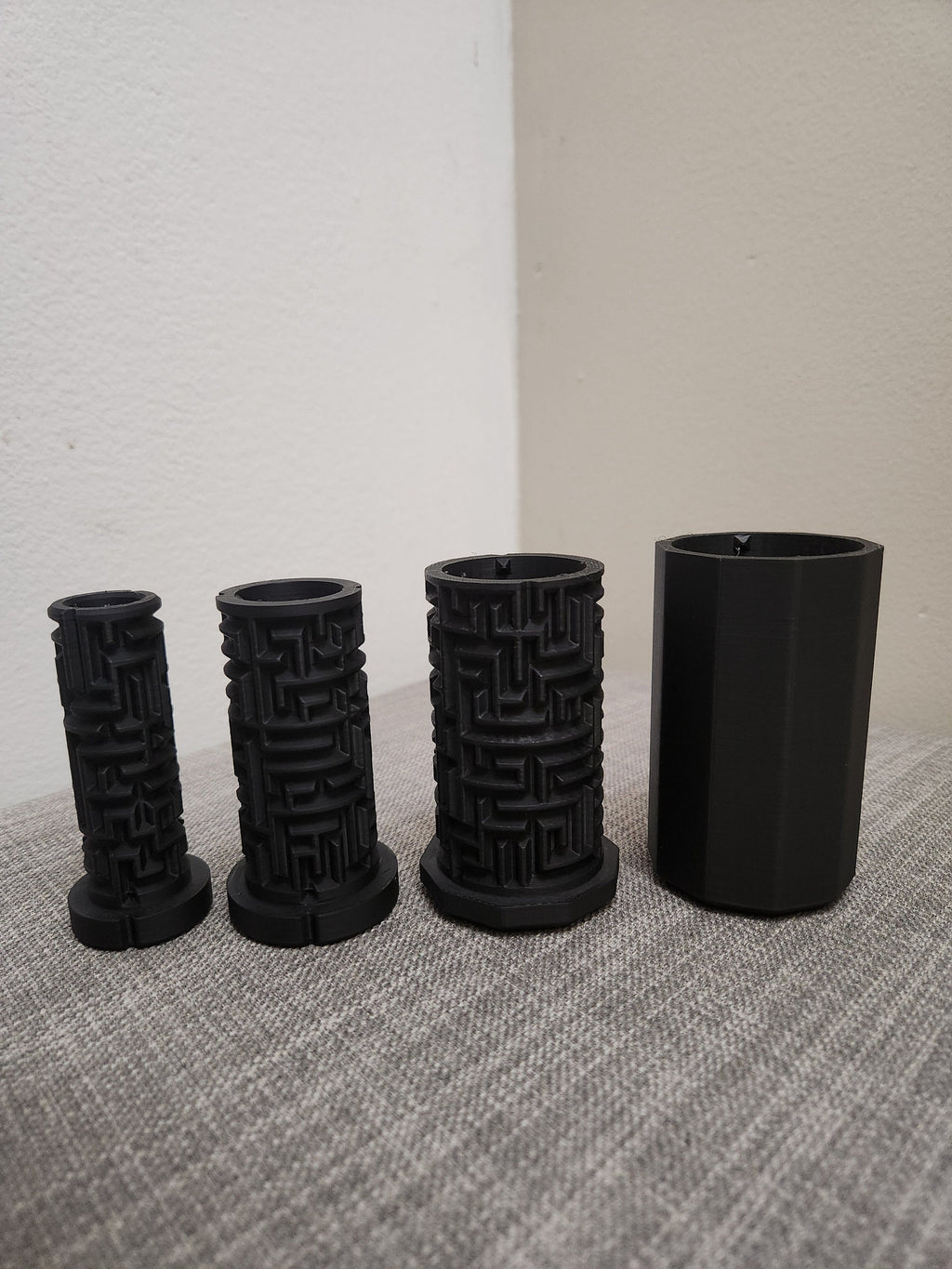 Four black 3D printed cylindrical objects, three with maze patterns, on a gray fabric surface.