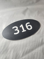 Oval black room number sign with white 316 text on textured surface