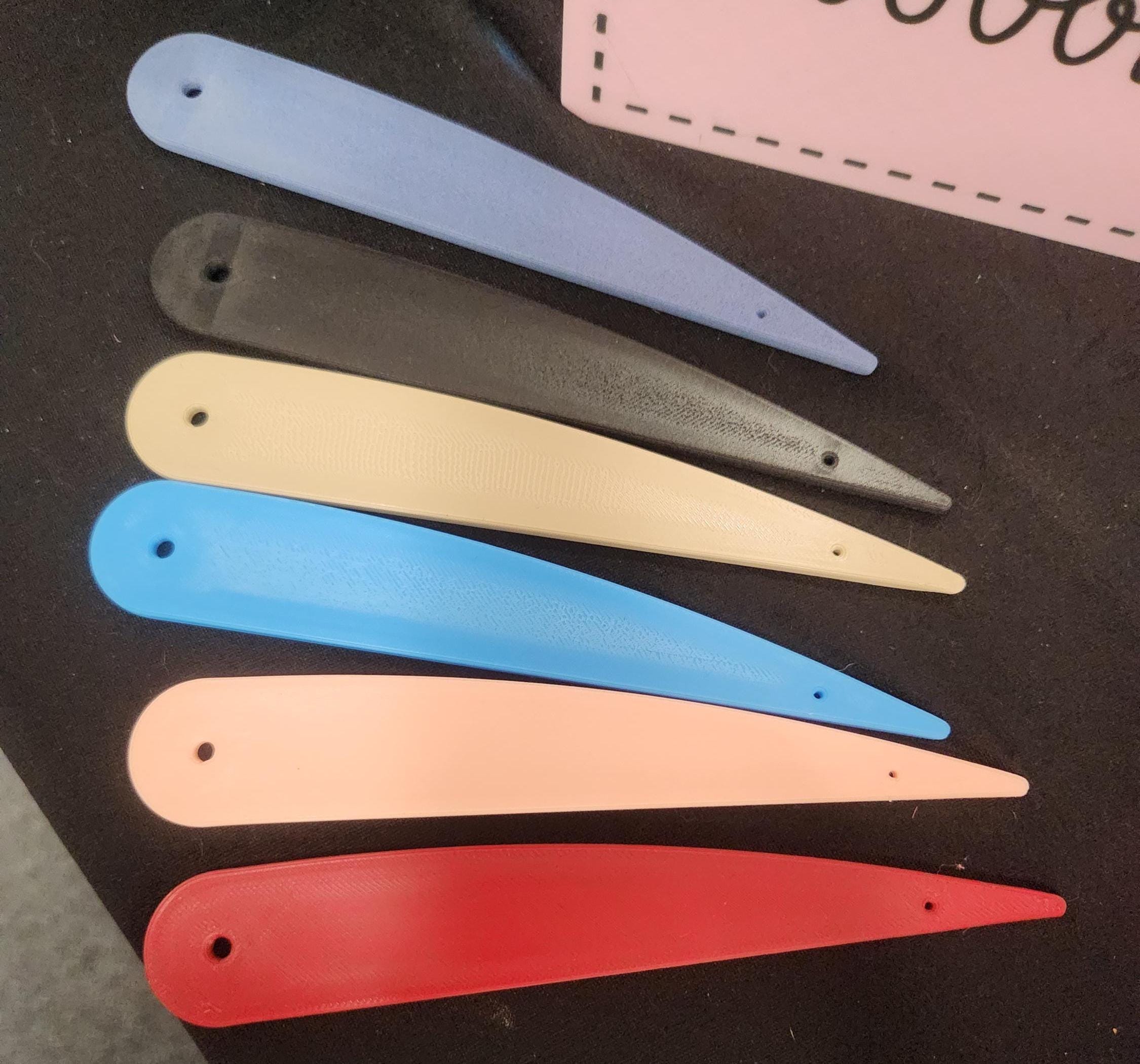 Assorted 3D printed plastic bookmarks in blue, black, beige, pink, and red on a black surface