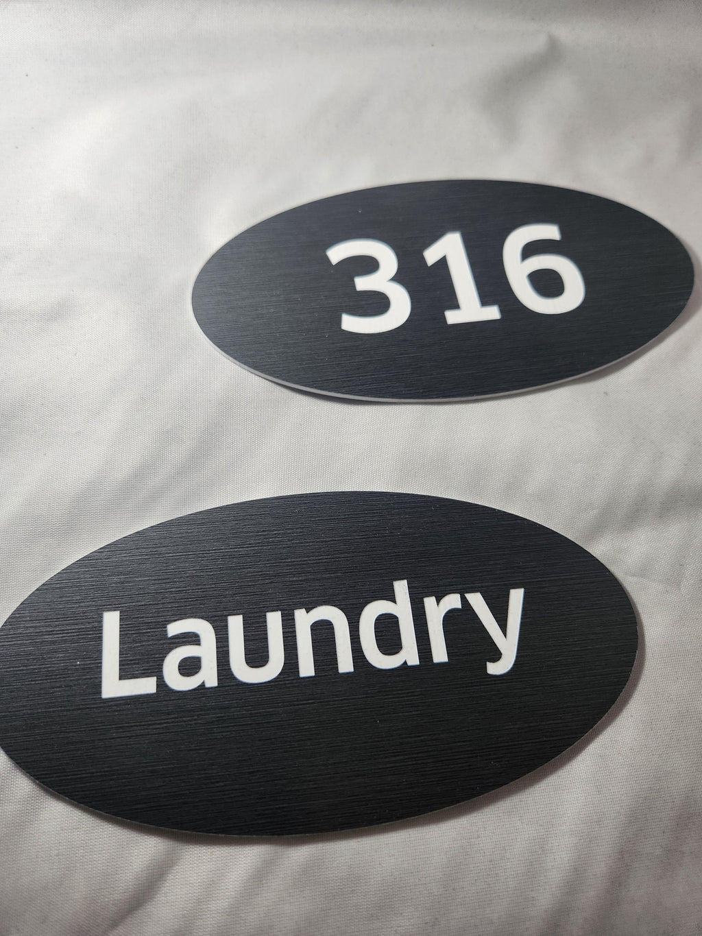 Oval black engraved signs with white text reading '316' and 'Laundry' on fabric background