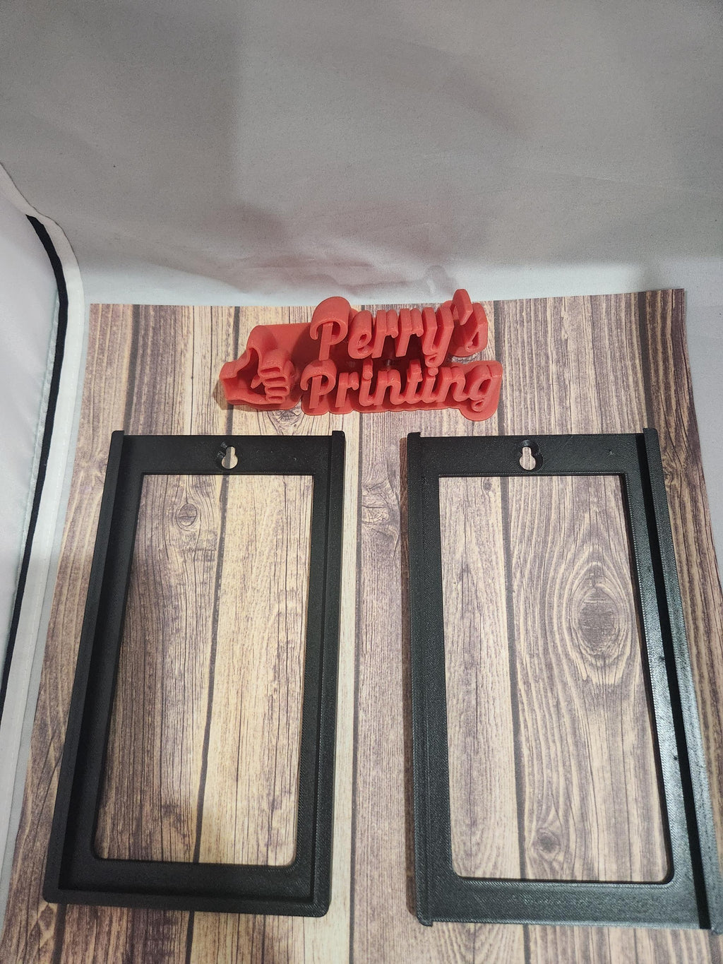 Two black 3D printed sign frames and a red Perry's Printing logo on wood background