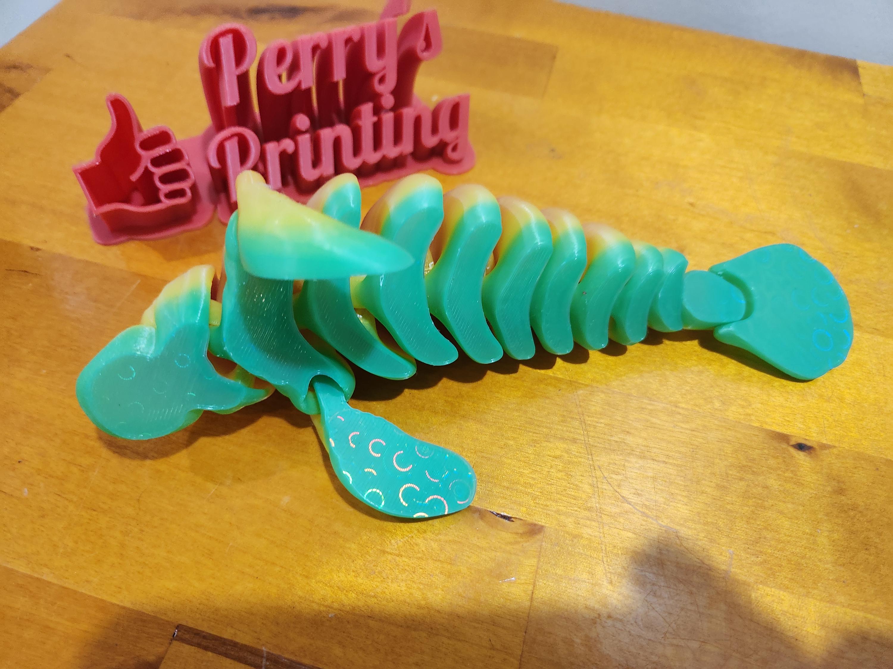 3D printed green bendable dinosaur figurine on wooden surface with Perry's Printing sign
