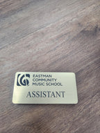 Custom engraved name tag reading 'Eastman Community Music School Assistant' on wood surface