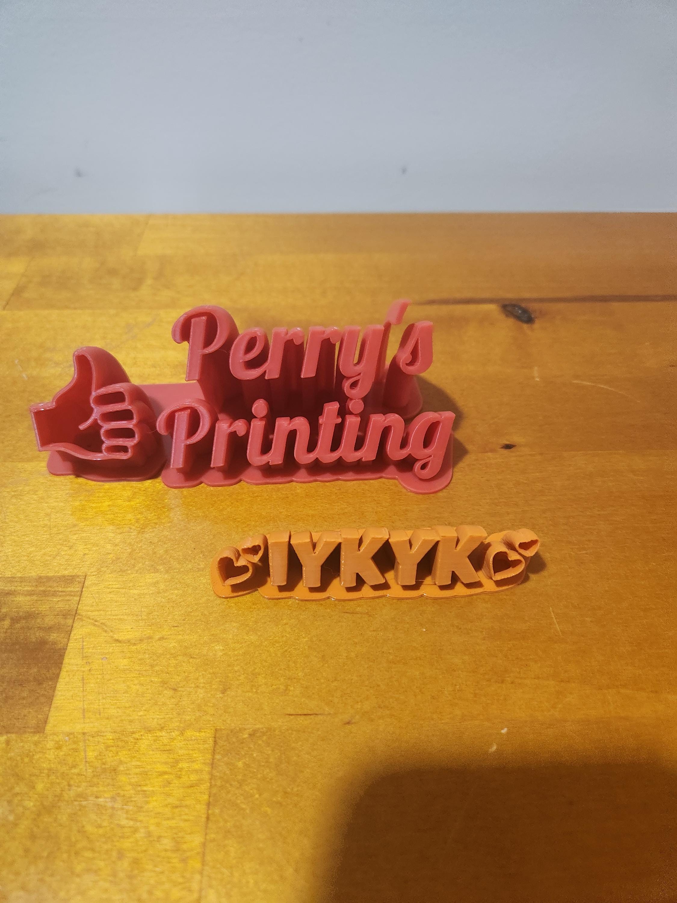 3D printed signs reading 'Perry's Printing' with thumbs up and 'IYKYK' with hearts on wood table