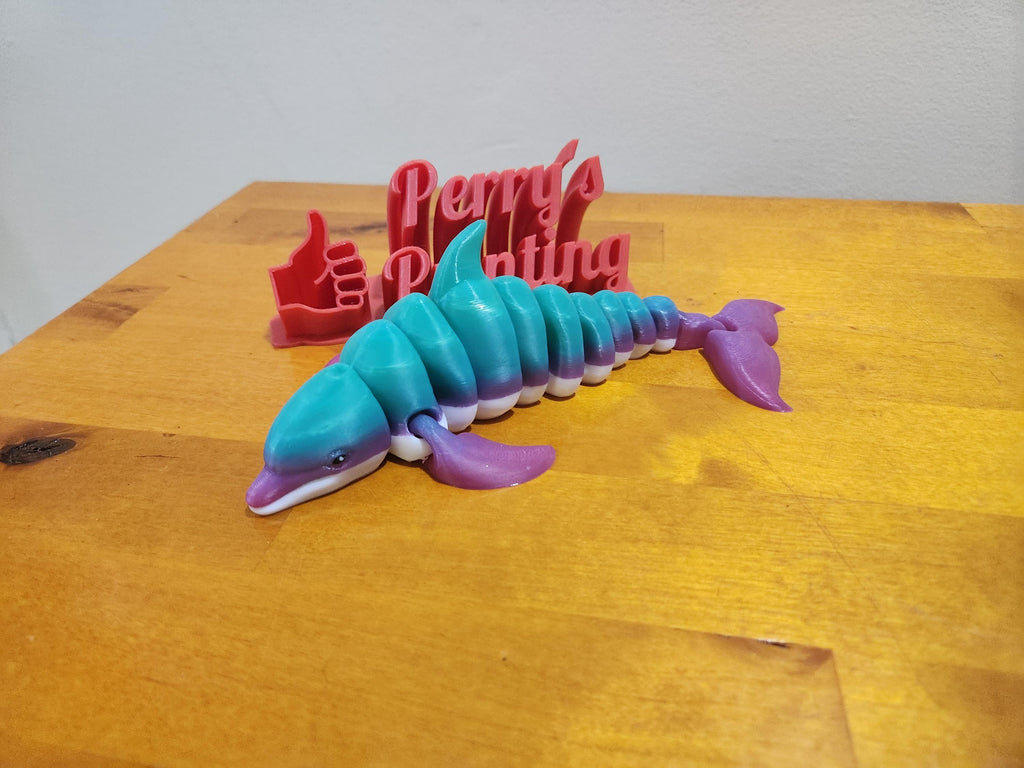 Colorful 3D printed articulated dolphin toy with red thumbs up sign on wooden table