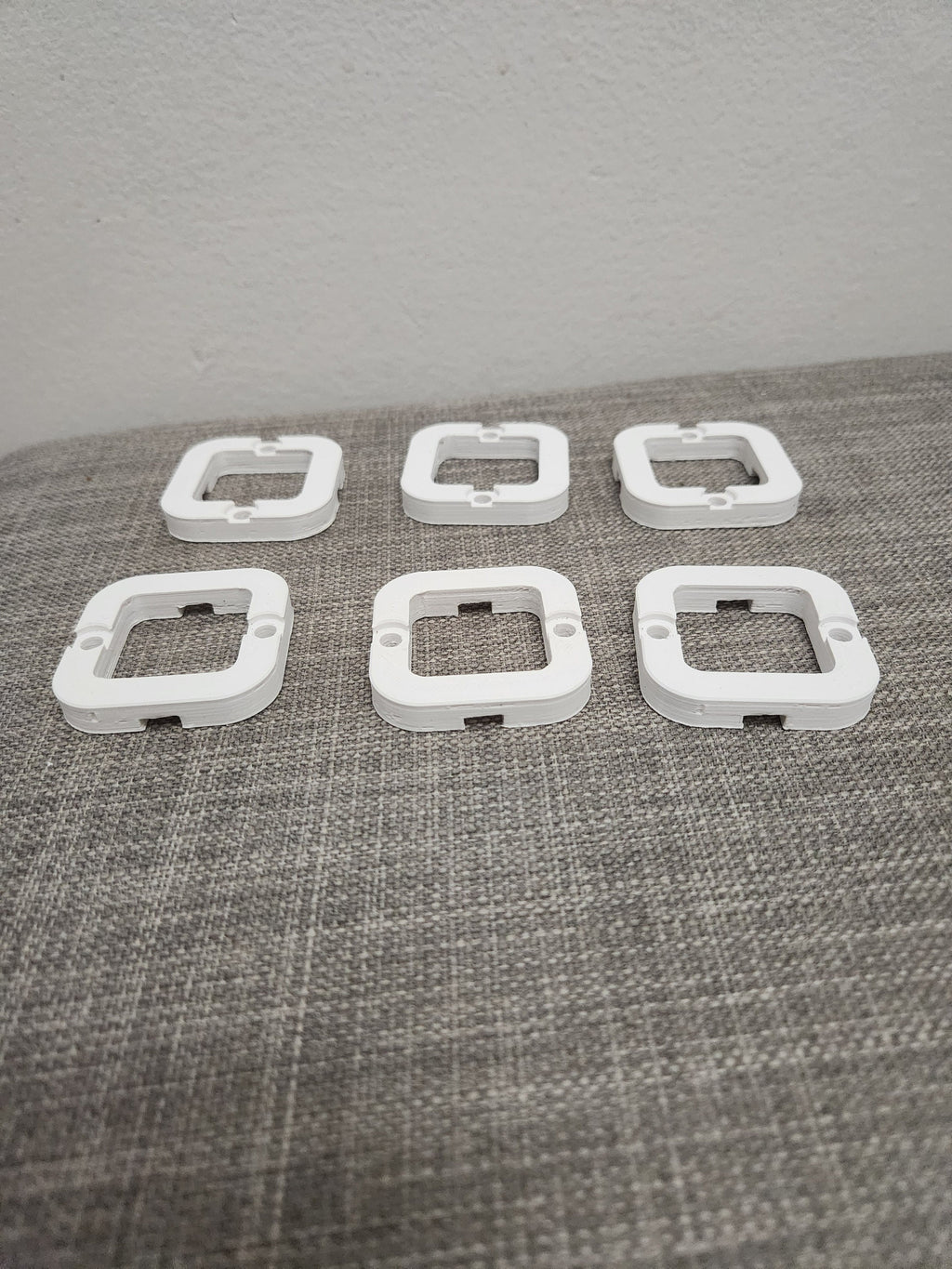 Six white 3D printed square plastic parts with screw holes on a gray fabric surface.