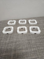 Six white 3D printed square plastic parts with screw holes on a gray fabric surface.