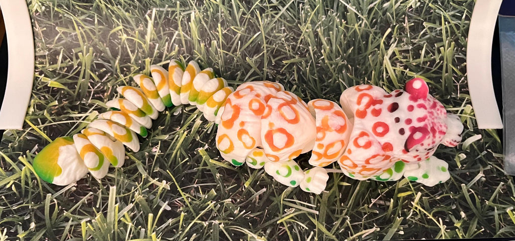 Colorful 3D printed articulated lizard with orange, green, and pink spots on grass background