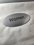 Brushed metal oval women restroom sign with black text on a light surface