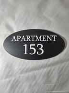 Custom oval black apartment number sign with white text reading 'Apartment 153'