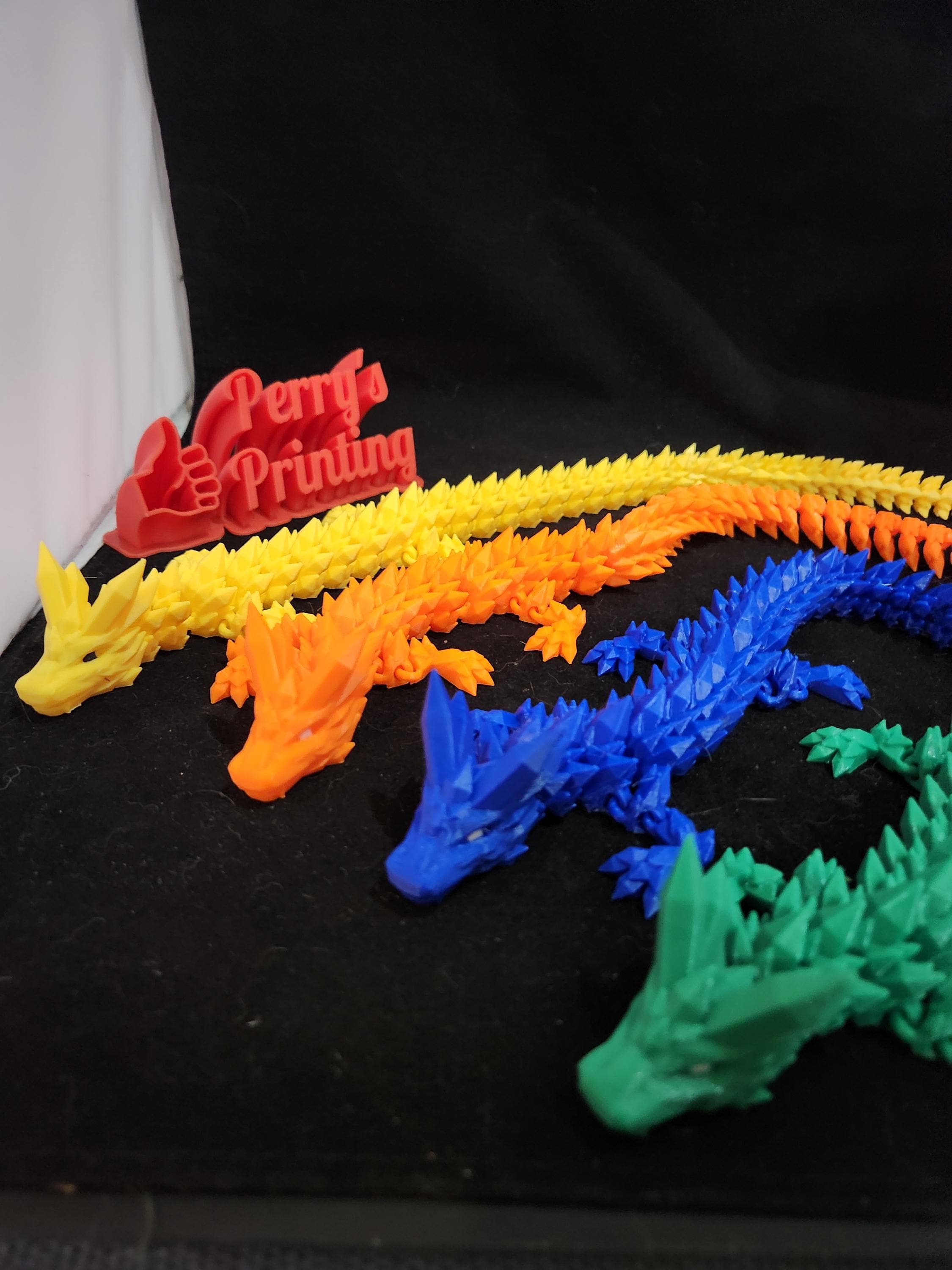 Colorful 3D printed articulated dragon toys in yellow, orange, blue, and green on black surface