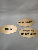 Gold oval engraved signs for office, restrooms, and employees only on gray background