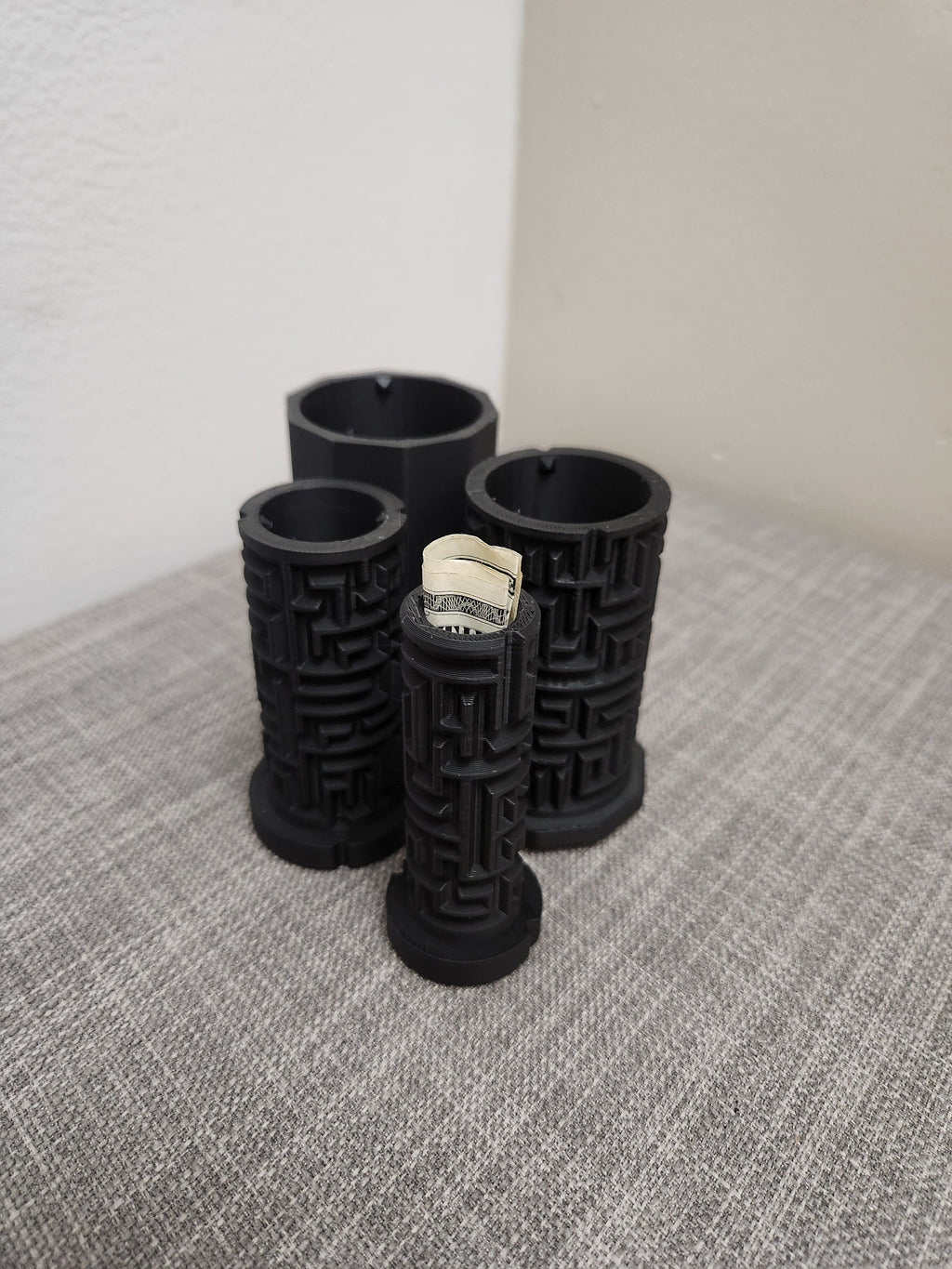 3D printed black puzzle tubes with geometric patterns, one holding rolled cash, on a table