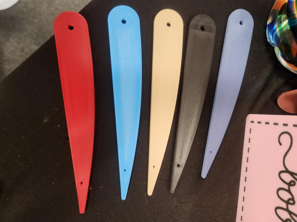 Set of five 3D printed plastic bookmarks in red, blue, cream, black, and light purple on black fabric.