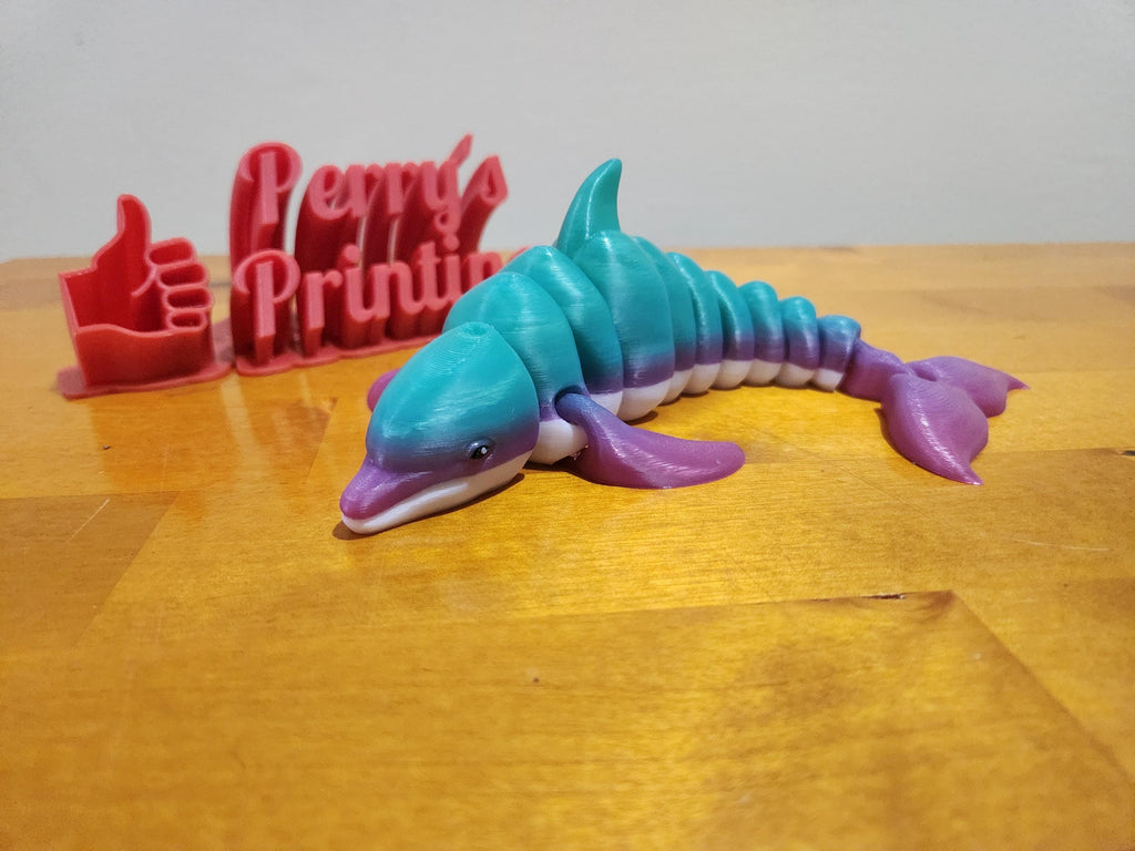 Colorful 3D printed dolphin figure on wood table, red thumbs-up sign in background