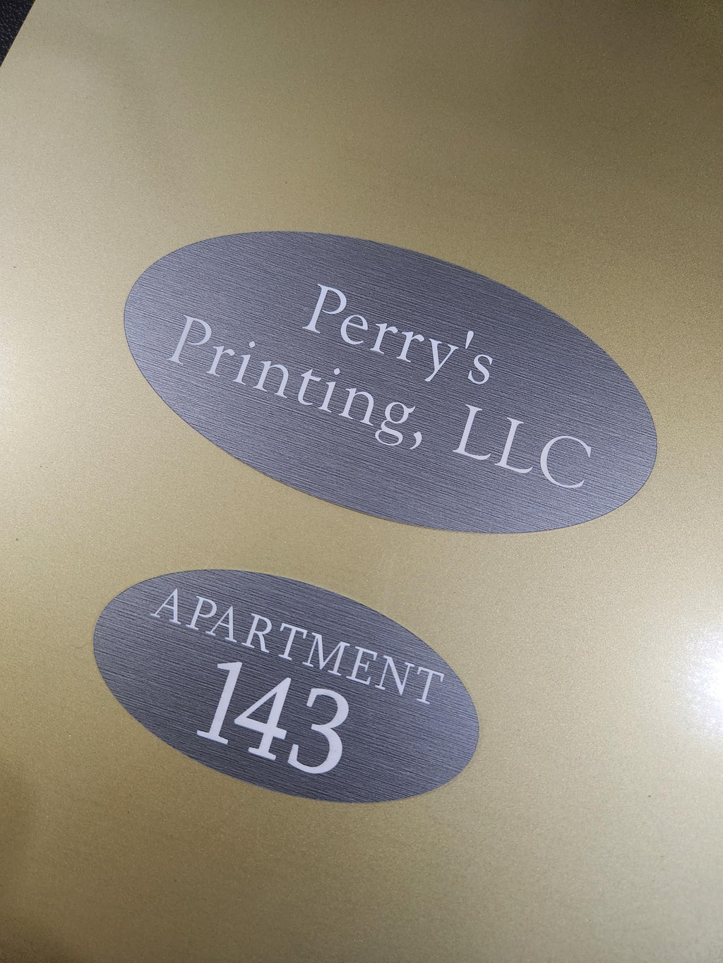 Custom gold door sign with gray ovals reading 'Perry's Printing, LLC' and 'Apartment 143'.