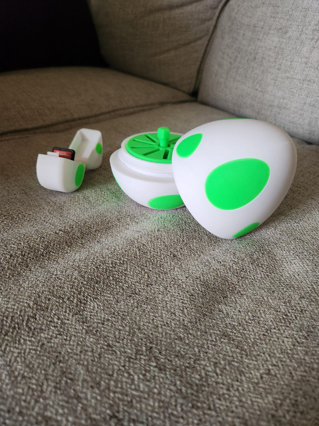 3D printed white egg with green spots, open to reveal Nintendo Switch game card holder, on a sofa.