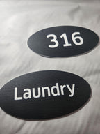 Custom black oval door signs with white text for room 316 and laundry, laser engraved.