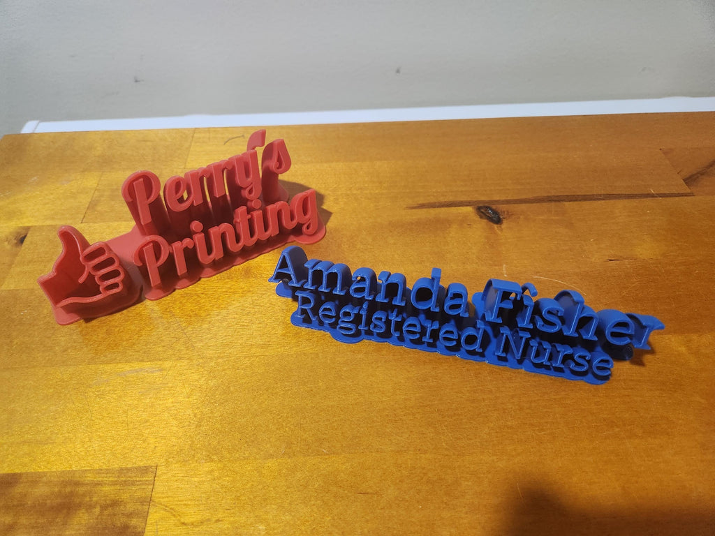 3D printed custom name signs in red and blue on a wooden surface, one with a thumbs up icon