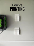 Two 3D printed wall mounts installed on a light gray wall, next to a green battery charger.