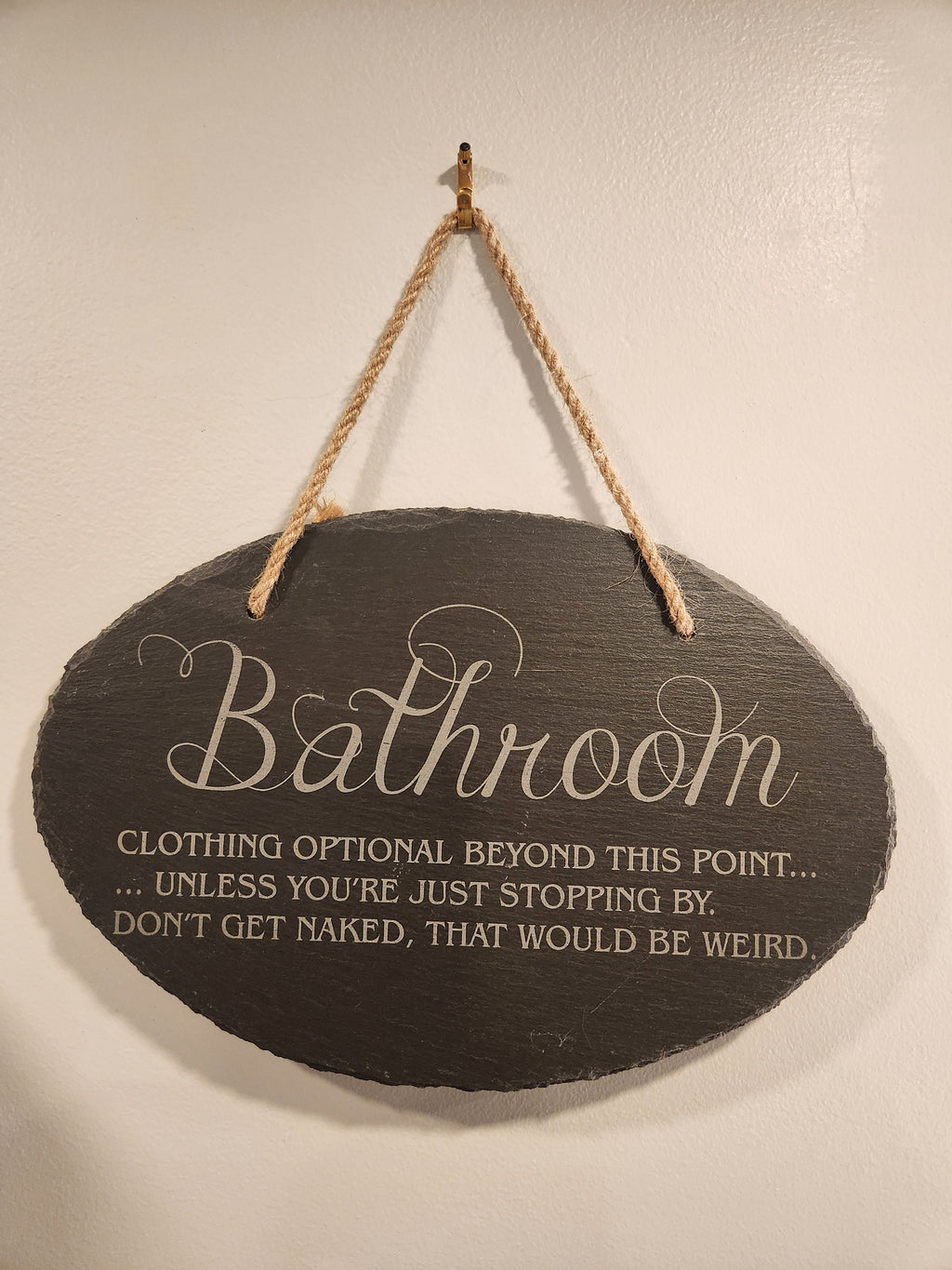 Engraved bathroom sign with humorous message hanging on a wall by a rope.