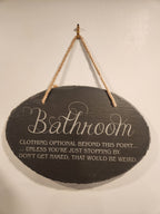 Engraved bathroom sign with humorous message hanging on a wall by a rope.