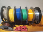 Multiple spools of colorful 3D printer filament on a tabletop organizer with a 3D printed sign