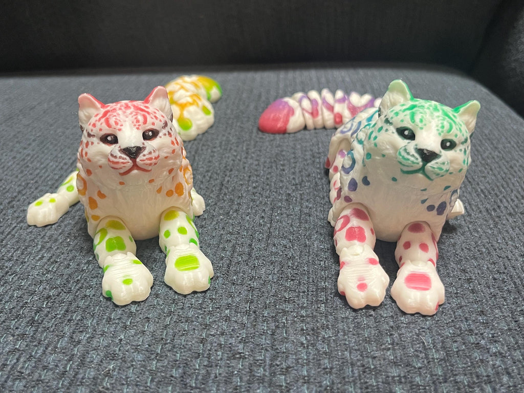 Colorful 3D printed articulated cat figurines with spotted patterns on a gray fabric surface
