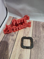 Red 3D printed Perry's Printing sign and small black square component on wood surface