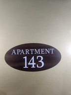 Oval woodgrain apartment number plaque with white 'Apartment 143' text on gold background