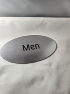 Brushed metal oval men’s restroom sign with black text on a white background.