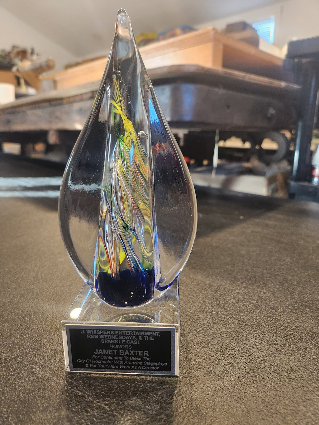 Glass teardrop-shaped award with colorful swirls on a plaque, displayed on a table.