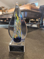 Glass teardrop-shaped award with colorful swirls on a plaque, displayed on a table.