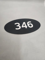 Black oval custom engraved sign with white number 346 on a light background