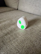 White 3D printed object with two green dots on a beige fabric sofa.