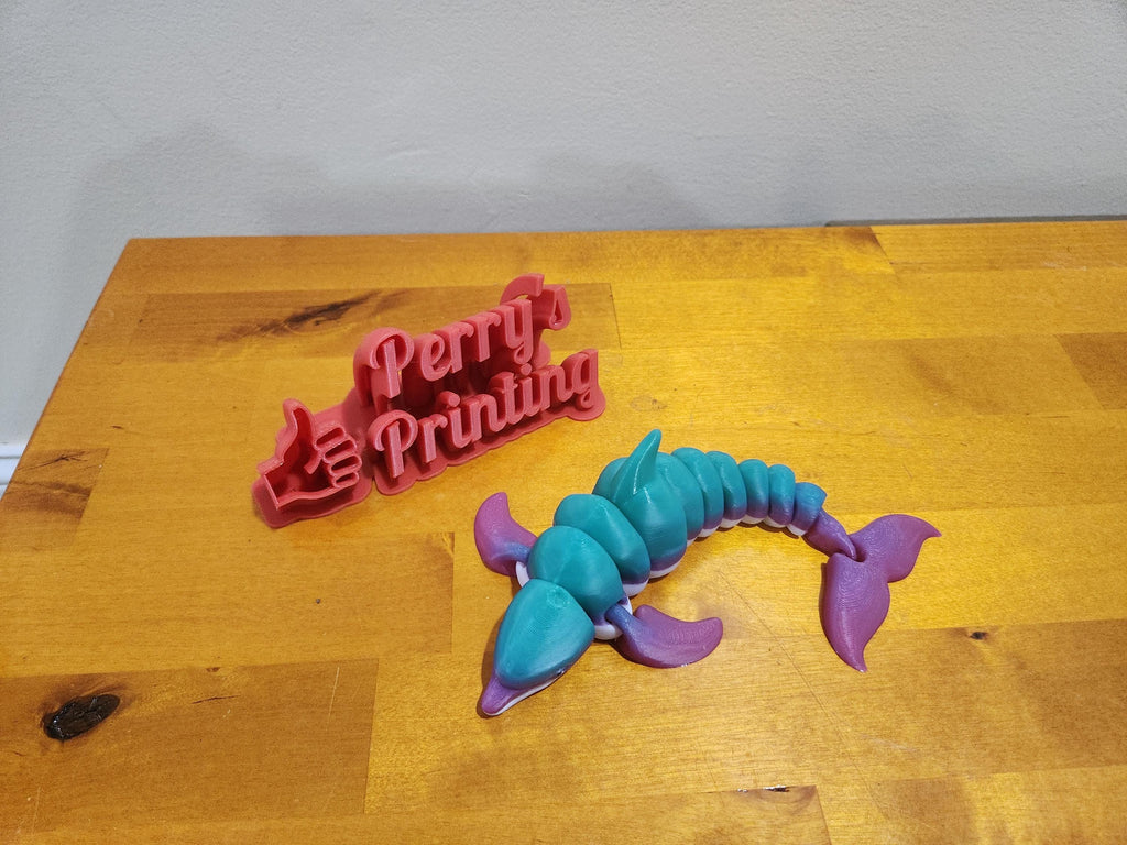 Colorful 3D printed articulated sea creature and red 'Perry's Printing' sign on wood table