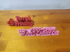 3D printed desk signs on wood surface, one reads 'Perry's Printing' with a thumbs up, the other 'Keep Calm and Carry On' with a crown.