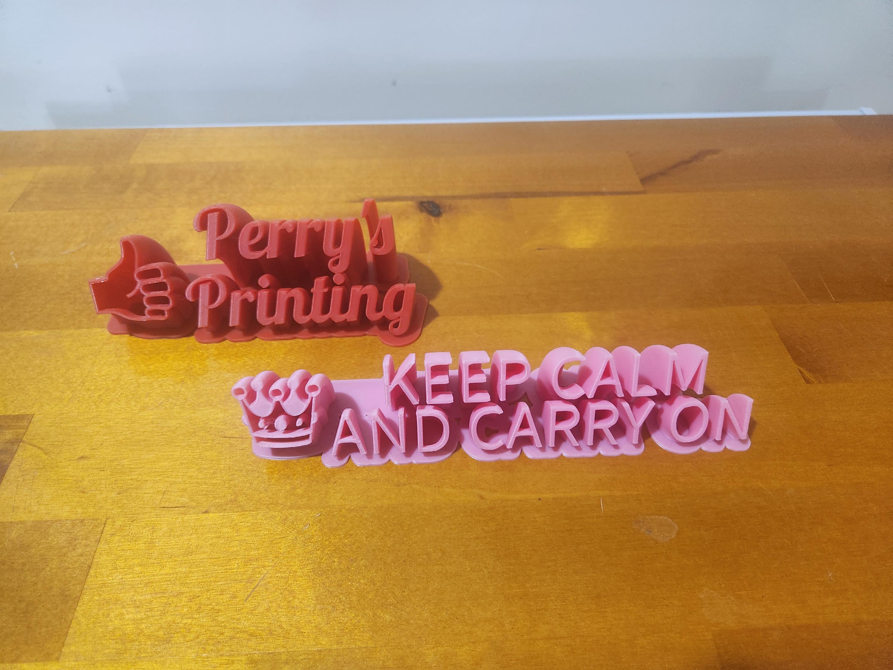 3D printed desk signs on wood surface, one reads 'Perry's Printing' with a thumbs up, the other 'Keep Calm and Carry On' with a crown.