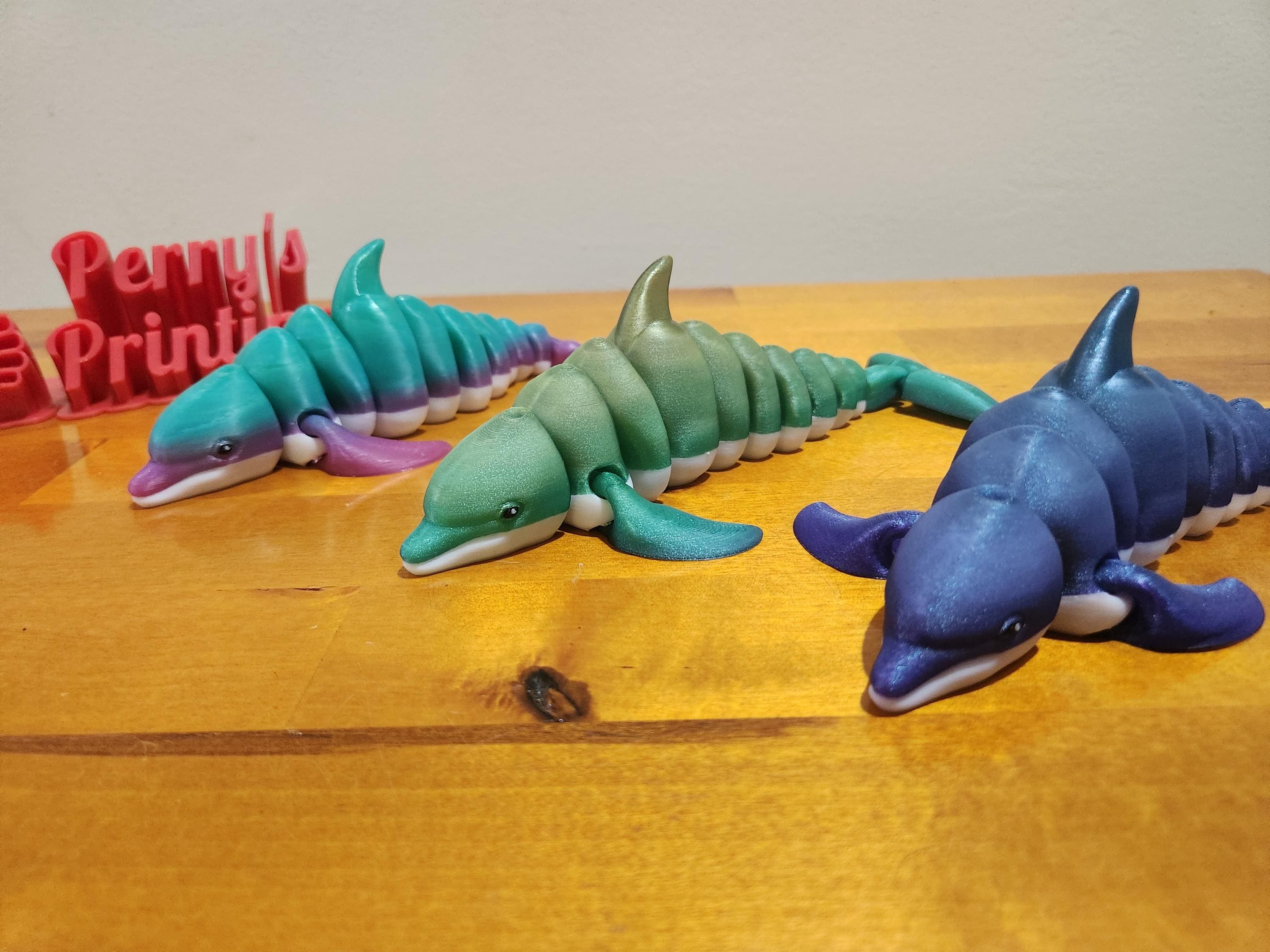 Three colorful 3D printed articulated dolphin toys displayed on a wooden surface