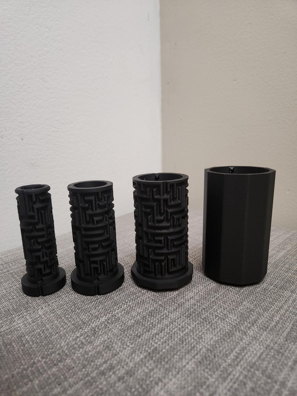 Four black 3D printed cylindrical objects with intricate maze designs on a grey fabric surface.