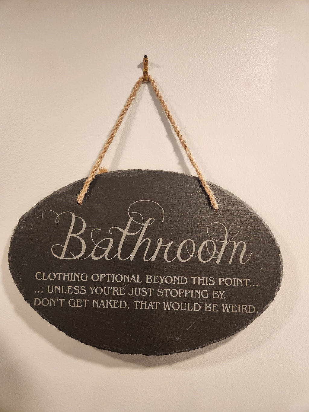 Funny engraved bathroom sign with humorous text, hanging on a wall with a rope.