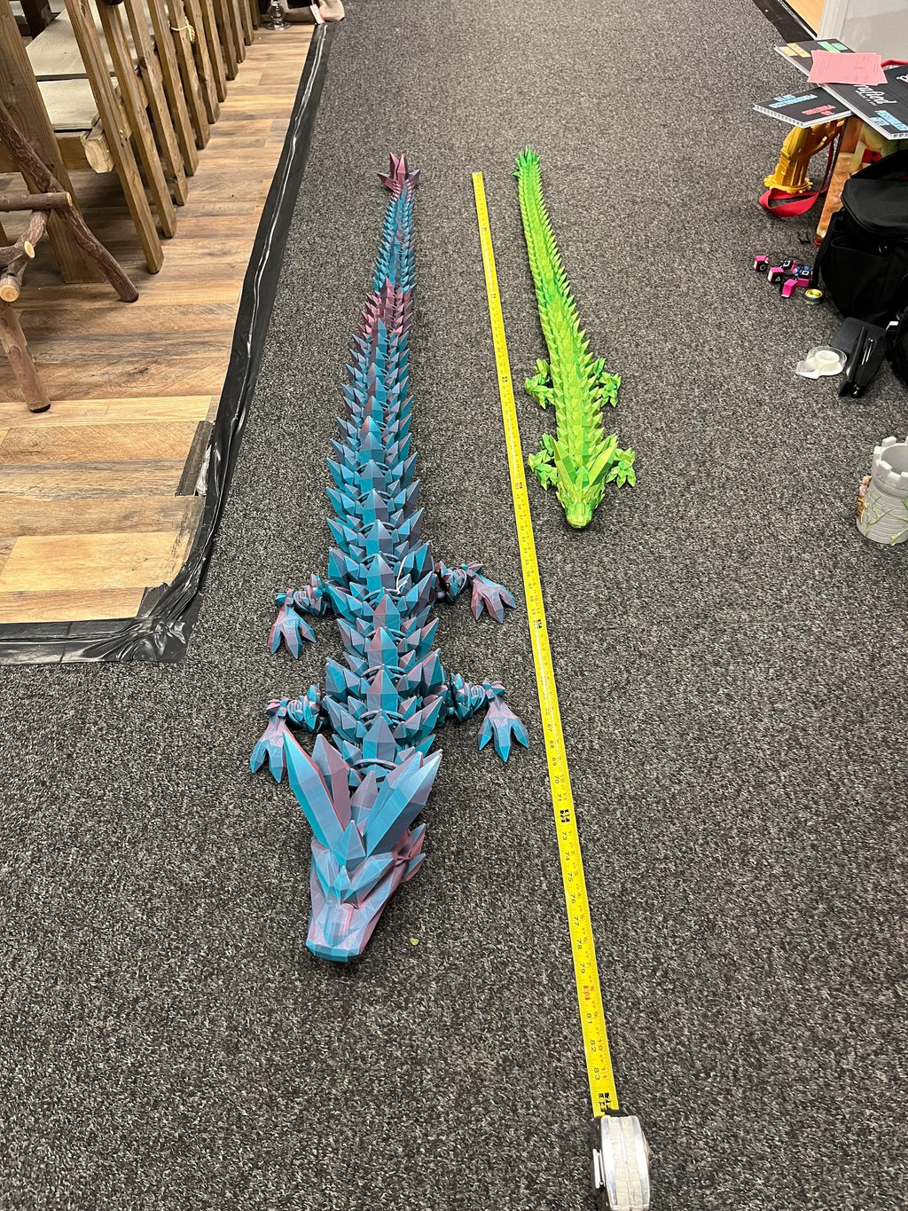 Two large articulated 3D printed dragon models, blue and green, beside a measuring tape on carpet
